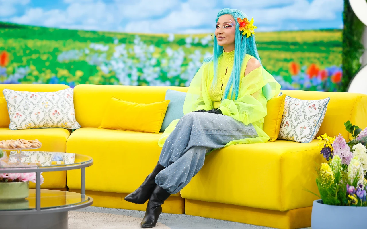Cleo w "halo tu polsat" kobieta z długimi turkusowymi włosami, ubrana w neonowy, limonkowy top i szare, luźne jeansy, siedzi na jaskrawo żółtej kanapie w studiu telewizyjnym z kolorową, kwiatową dekoracją; w tle panorama łąki pełnej kwiatów