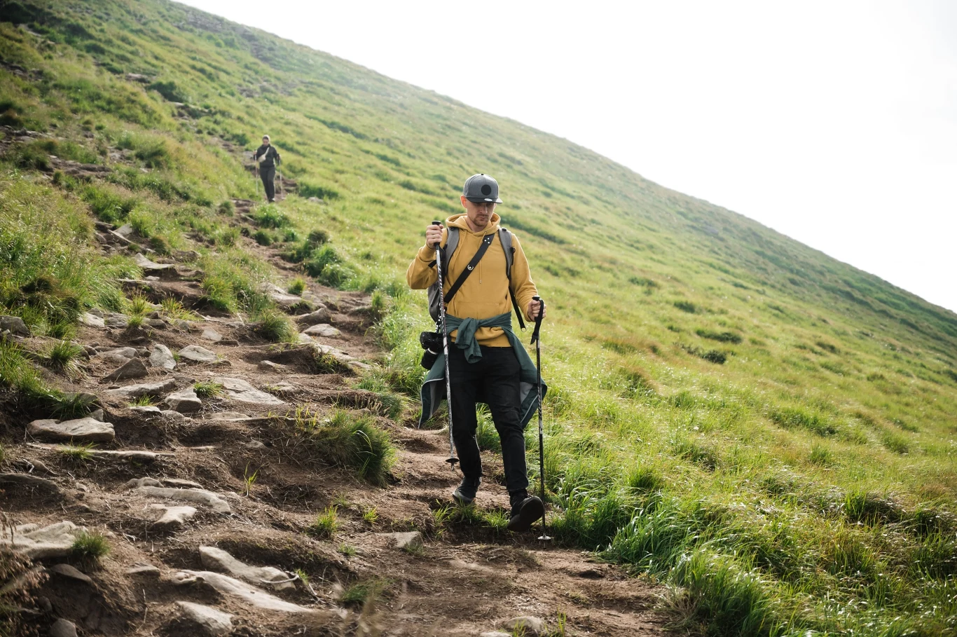 mężczyzna w żółtej kurtce i z plecakiem schodzi kamienistą ścieżką po zboczu zielonego wzgórza, używając kijków trekkingowych; w tle kolejna osoba idzie w tym samym kierunku