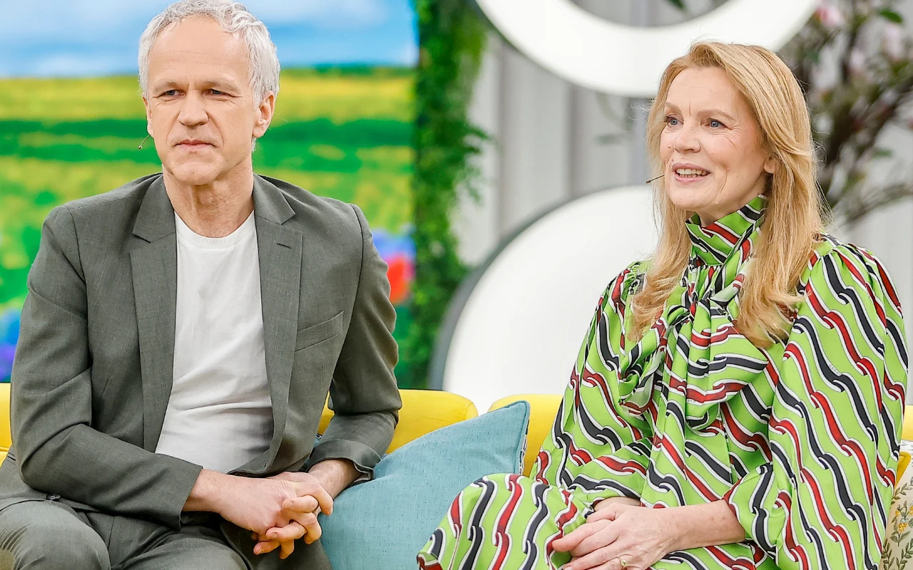 Razem na scenie i razem w domu Dwoje dorosłych siedzi obok siebie na żółtej sofie w jasnym, nowoczesnym wnętrzu; mężczyzna w szarym garniturze i białej koszulce ma jasne, krótkie włosy, a kobieta w kolorowej, wzorzystej sukni uśmiecha się i ma długie blond włosy, w tle widać zielony...