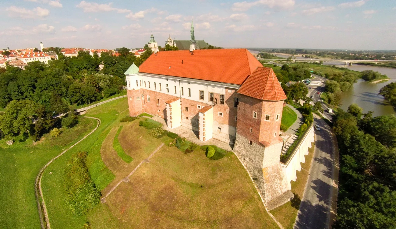Czy można zwiedzać zamek w Sandomierzu? Zamek w Sandomierzu z czerwonym dachem na wzgórzu, otoczony zielenią, z widoczną rzeką po prawej stronie