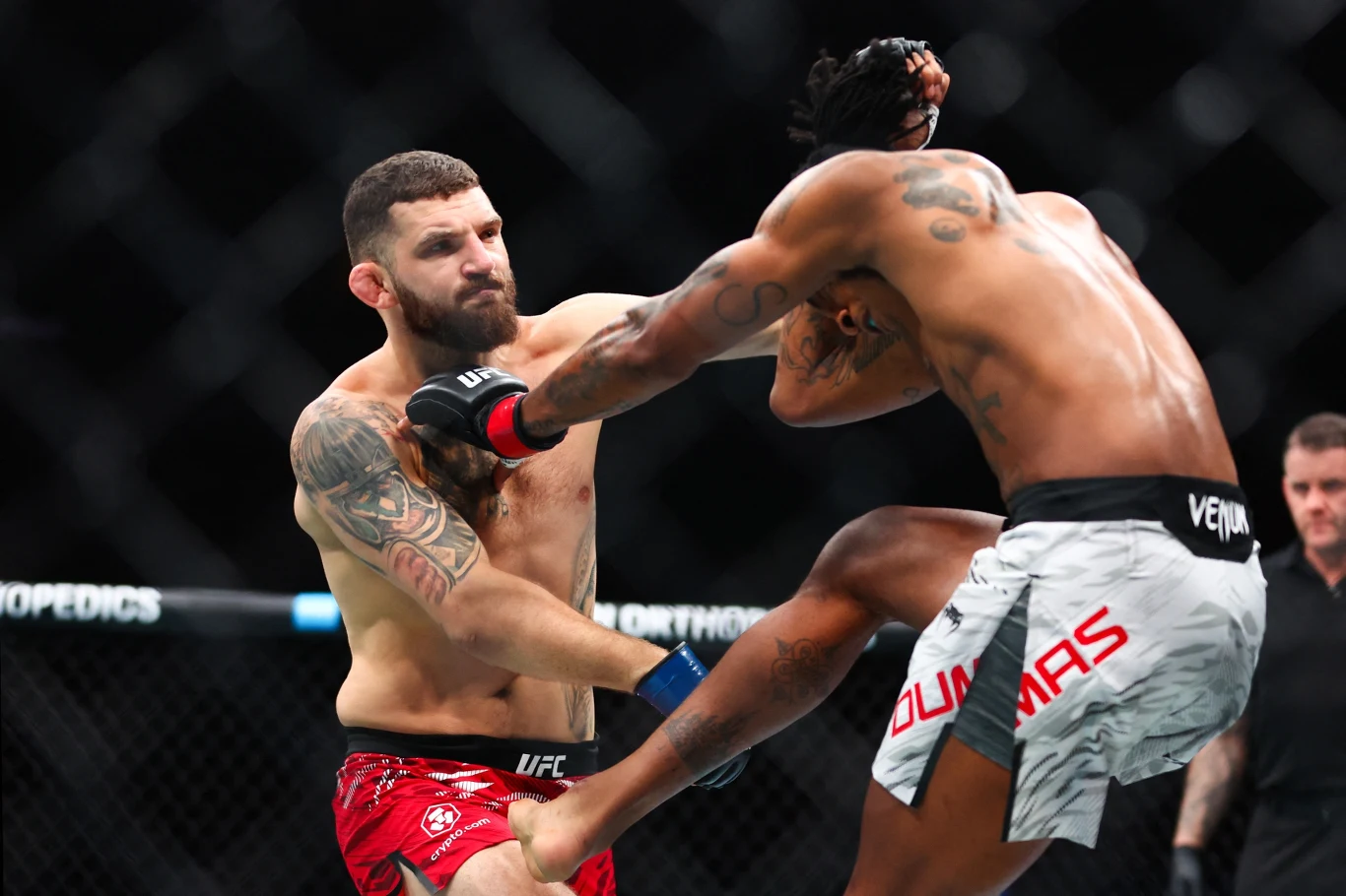 Dwóch zawodników MMA w oktagonie podczas wymiany ciosów, jeden wykonuje wysoki kopniak w stronę głowy przeciwnika, który próbuje się bronić, widoczne napięcie mięśniowe i intensywna akcja sportowa.