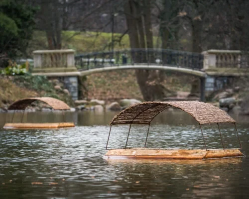 Drewniane tratwy z zadaszeniami z gałęzi unoszą się na spokojnej tafli wody, w tle widoczny jest kamienny mostek otoczony drzewami.