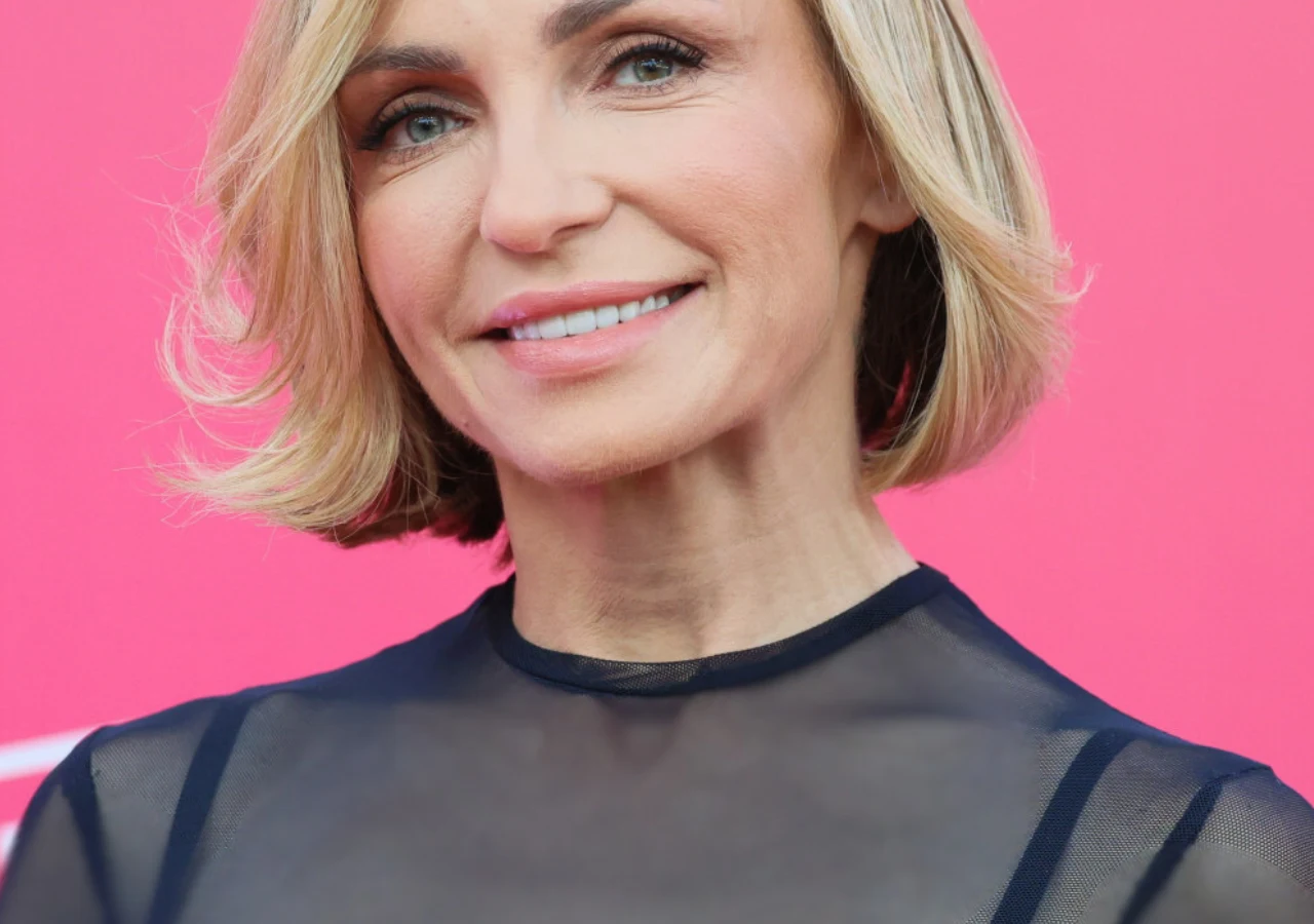 Anna Wyszkoni Anna Wyszkoni, blond włosy do ramion w falach, czarna półtransparentna bluzka, różowe tło