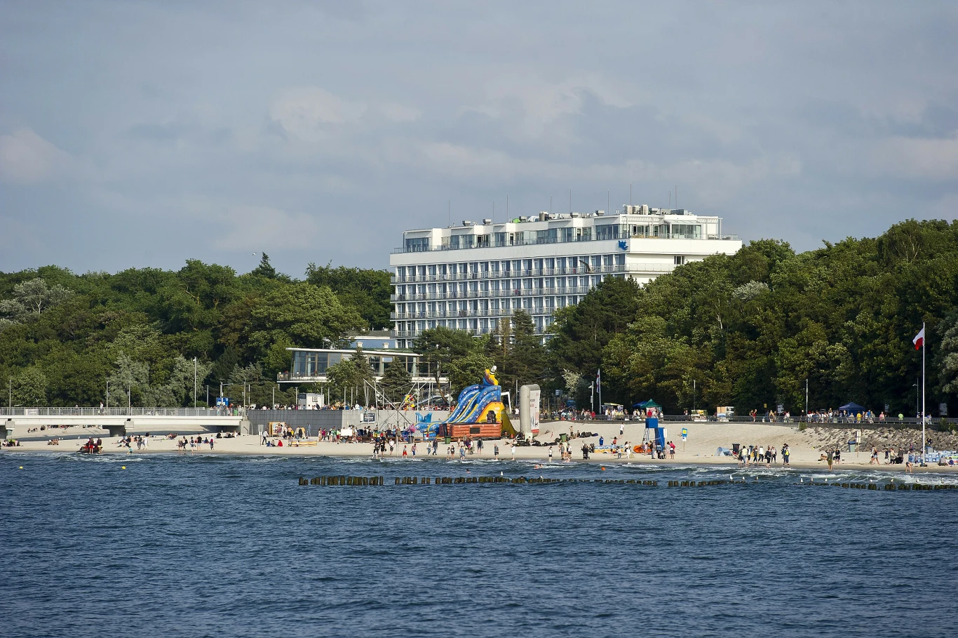 biały, nowoczesny hotel w otoczeniu zielonych drzew położony tuż przy szerokiej piaszczystej plaży z tłumem ludzi i kolorowymi atrakcjami dla dzieci, wszystko nad morzem