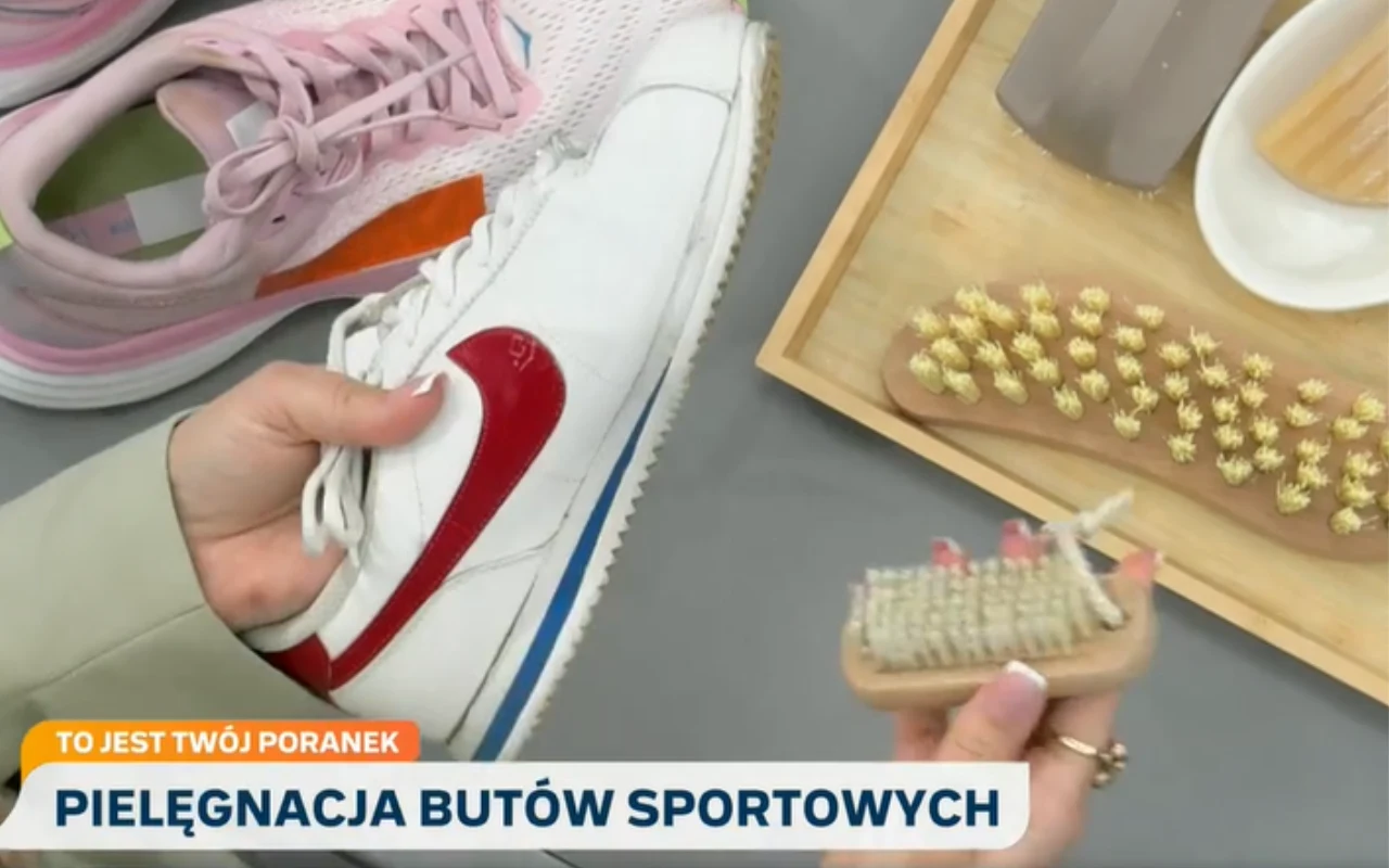 Pielęgnacja butów sportowych Biała sportowa sneakerka z czerwonym logo Nike trzymana w jednej ręce, w drugiej ręce szczotka do czyszczenia, obok na tacy inne szczotki oraz mydelniczka; w tle para różowych sportowych butów.