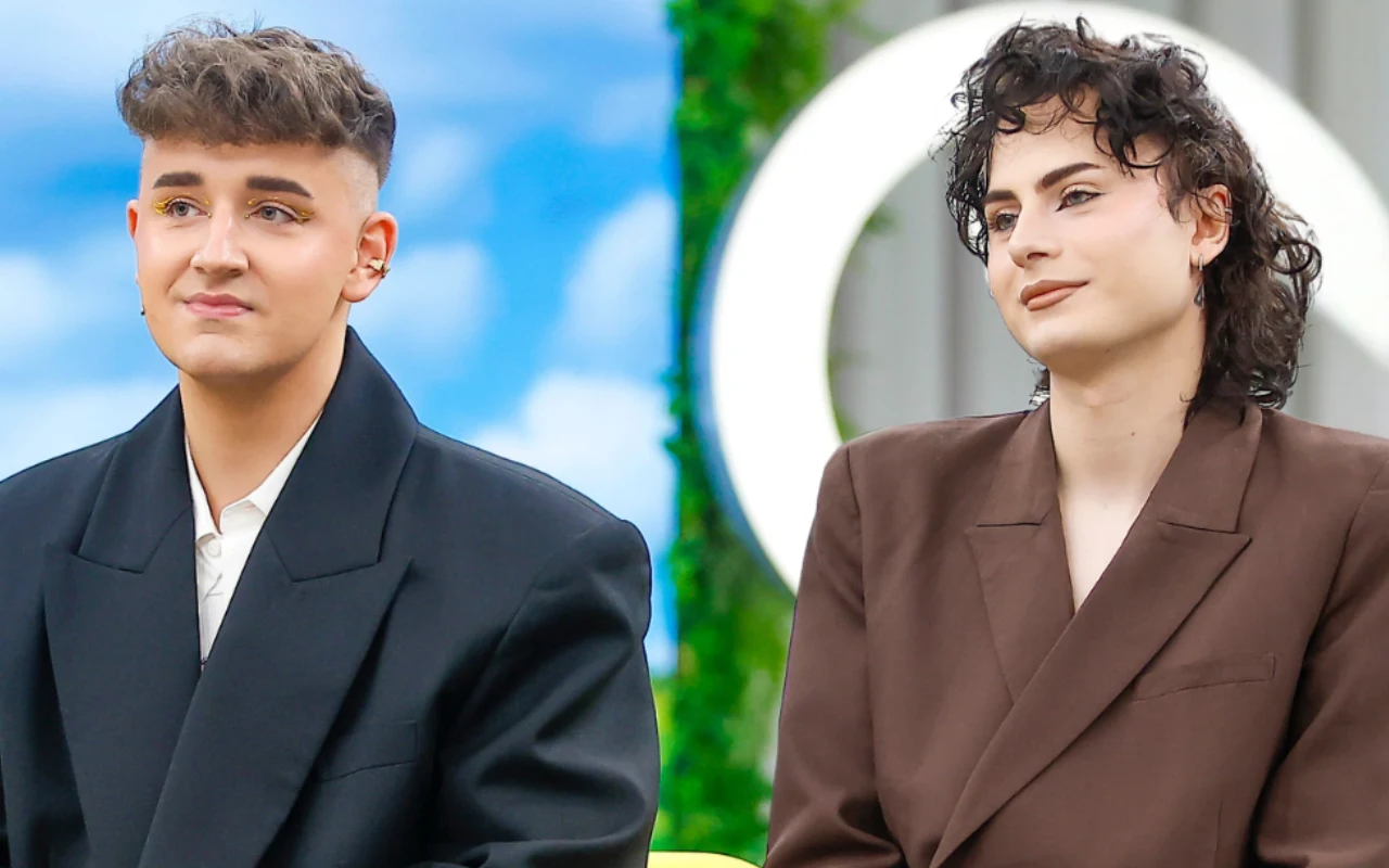 Dwoje młodych ludzi siedzi obok siebie, oboje ubrani w eleganckie, ciemne marynarki, z neutralnymi wyrazami twarzy; w tle widoczne są zielone rośliny i okrągły, biały element dekoracyjny.