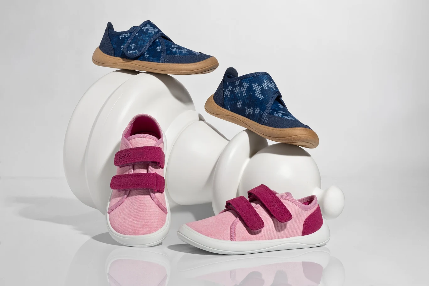 Dwa modele dziecięcych butów w pastelowych kolorach, para różowych z zapięciami na rzepy na pierwszym planie oraz dwie pary niebieskich z wzorem w psy i gumową podeszwą na tle dużej figurki szachowego pionka w odcieniu bieli.