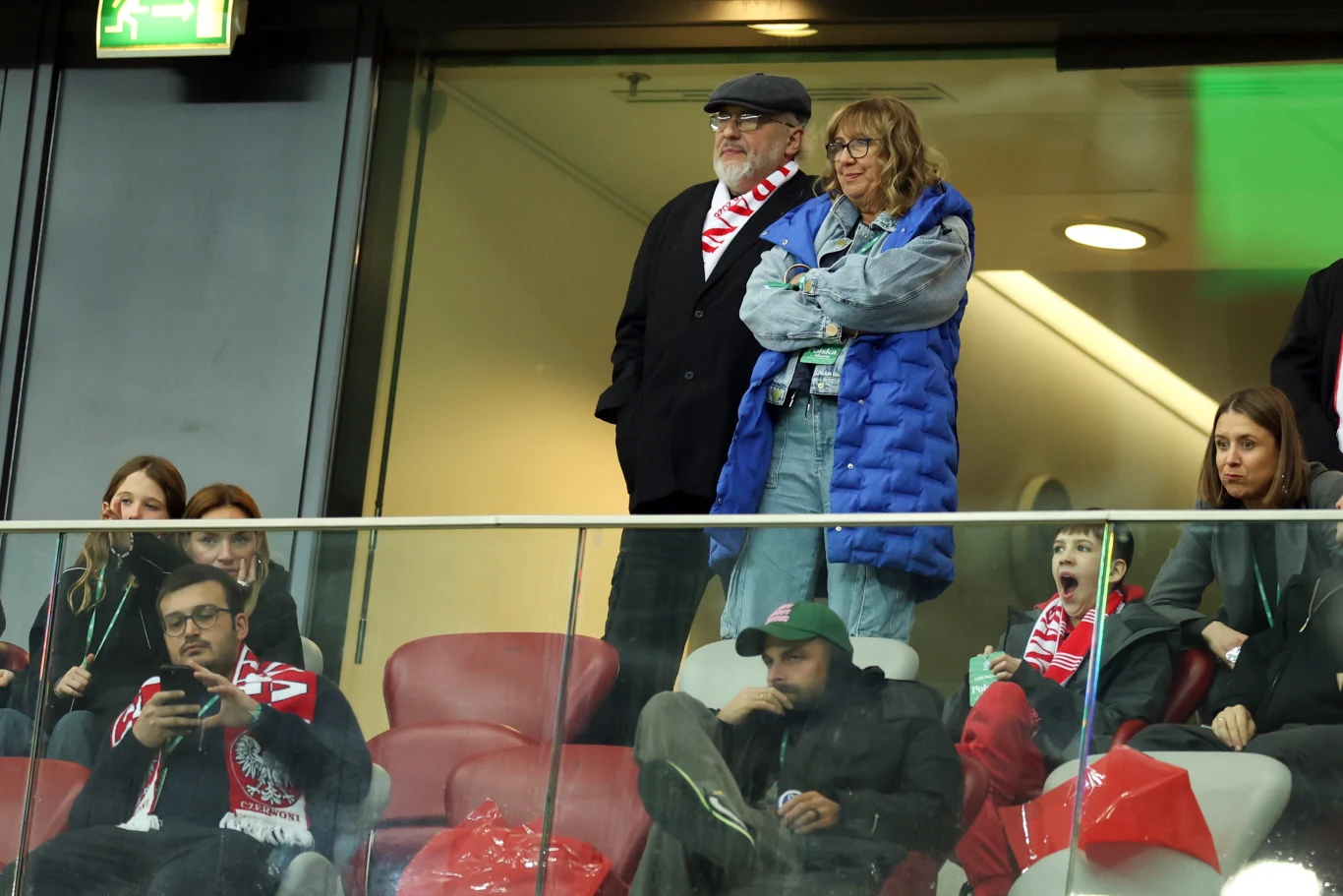 Grupa kibiców ubranych w szaliki i stroje w barwach narodowych obserwuje wydarzenia na stadionie, część siedzi, inni stoją, niektórzy okazują emocje, a atmosfera jest skupiona i lekko napięta.