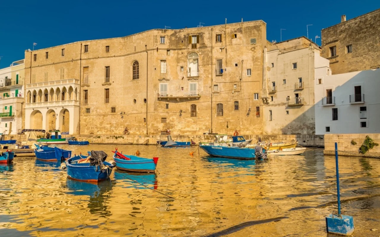 Zabytkowe kamienice o piaskowym kolorze odbijające się w spokojnej wodzie portu, wokół których cumują małe niebieskie i czerwone łodzie rybackie w promieniach słońca.