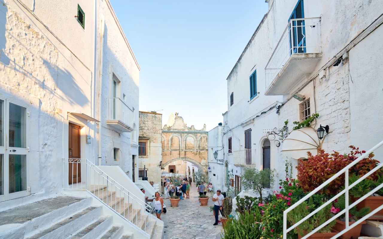 Wąska, brukowana uliczka otoczona bielonymi kamiennymi domami, liczne doniczki z roślinami przy schodach, w tle zabytkowa brama z łukami, spacerujący mieszkańcy i turyści w popołudniowym świetle.