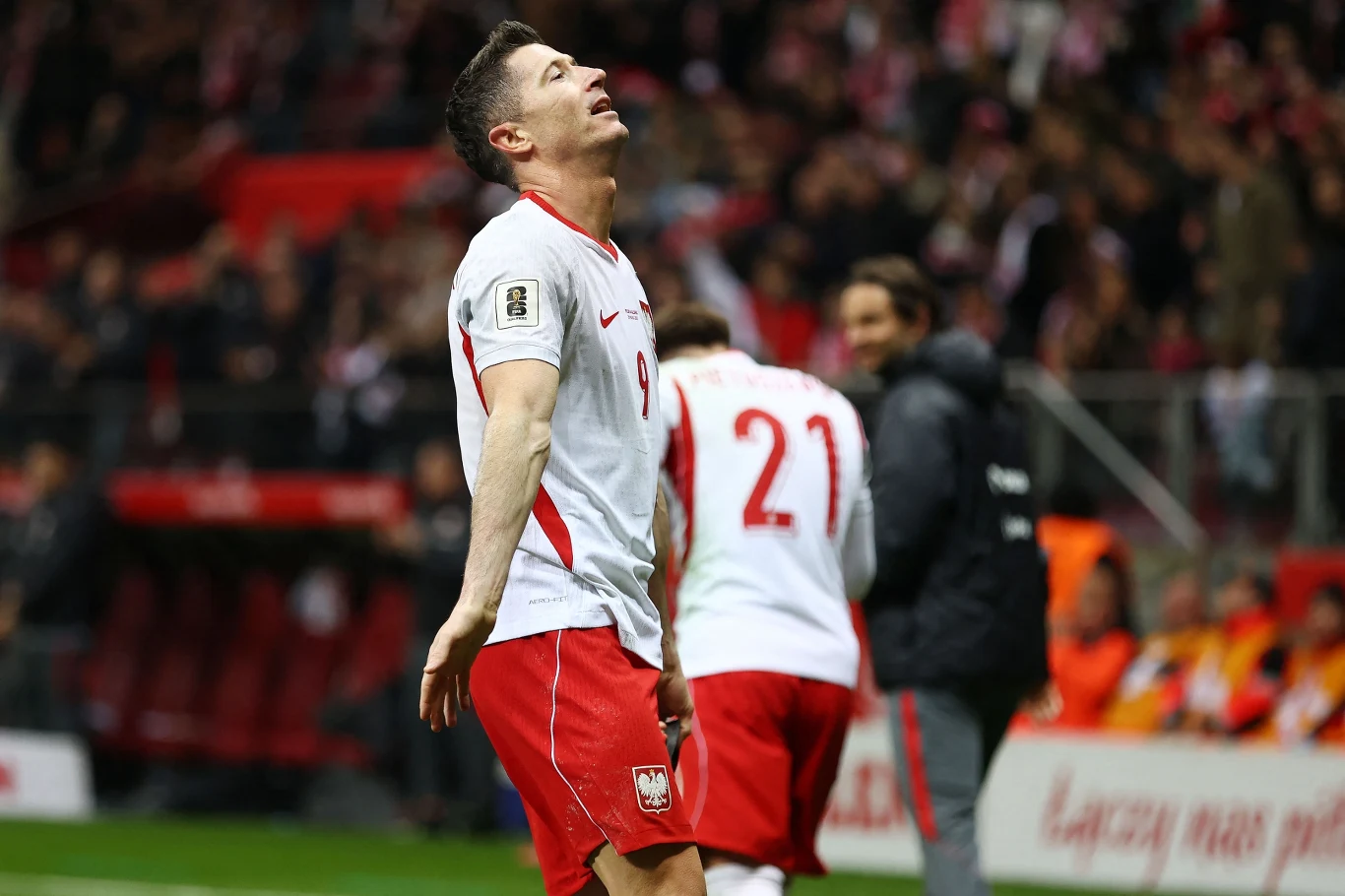 piłkarz w biało-czerwonym stroju z numerem 9 wyraża frustrację lub rozczarowanie na boisku, w tle widoczny zawodnik z numerem 21 oraz kibice na trybunach