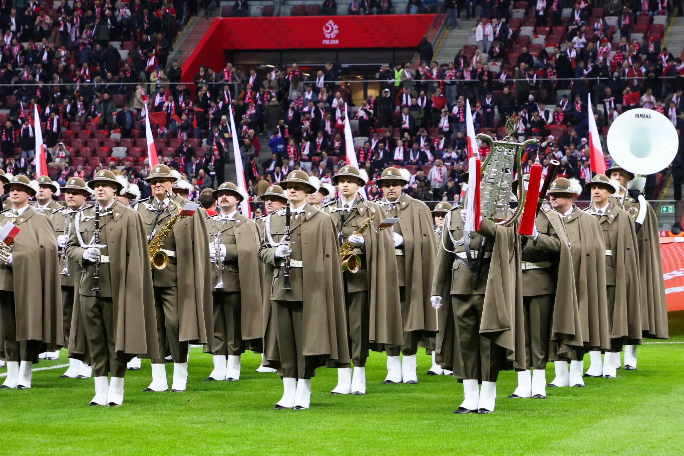orkiestra wojskowa w historycznych mundurach grająca na instrumentach dętych stoi w rzędach na zielonej murawie stadionu, w tle widoczni kibice z biało-czerwonymi szalikami