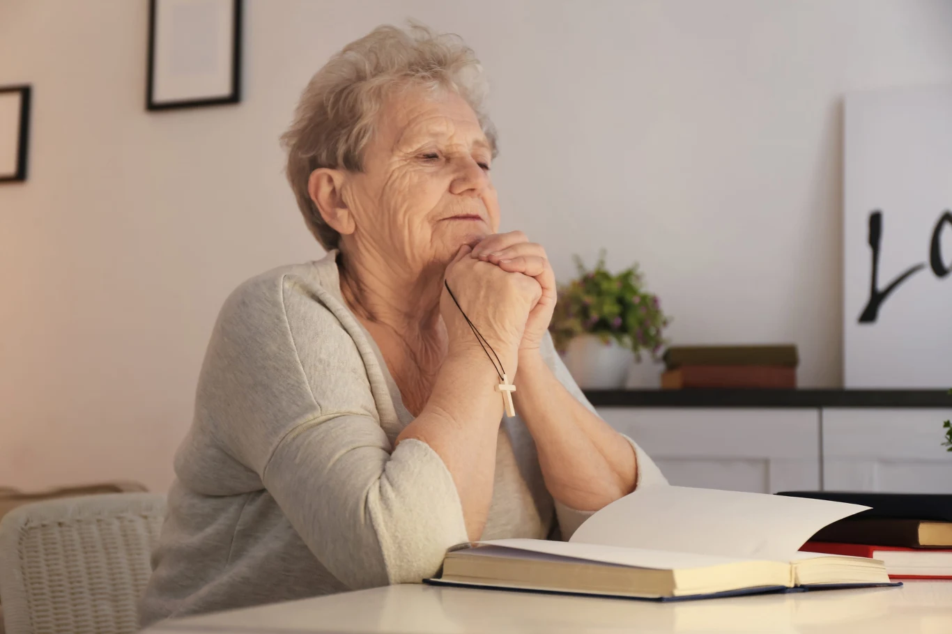 starsza kobieta siedząca przy stole z zamkniętymi oczami, trzymająca różaniec w dłoniach opartych o otwartą książkę, w tle kwiaty oraz ozdobne napisy na ścianie