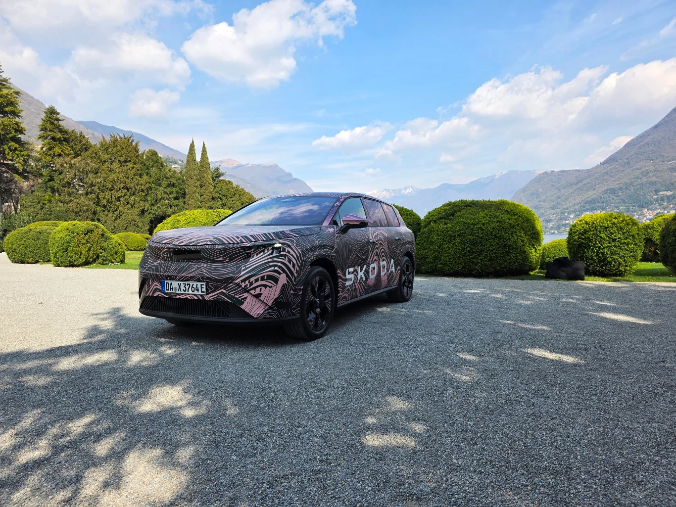 samochód Skoda w prototypowym kamuflażu zaparkowany na szerokiej alejce żwirowej, w otoczeniu przyciętych zielonych krzewów, z górzystym krajobrazem i częściowo zachmurzonym niebem w tle