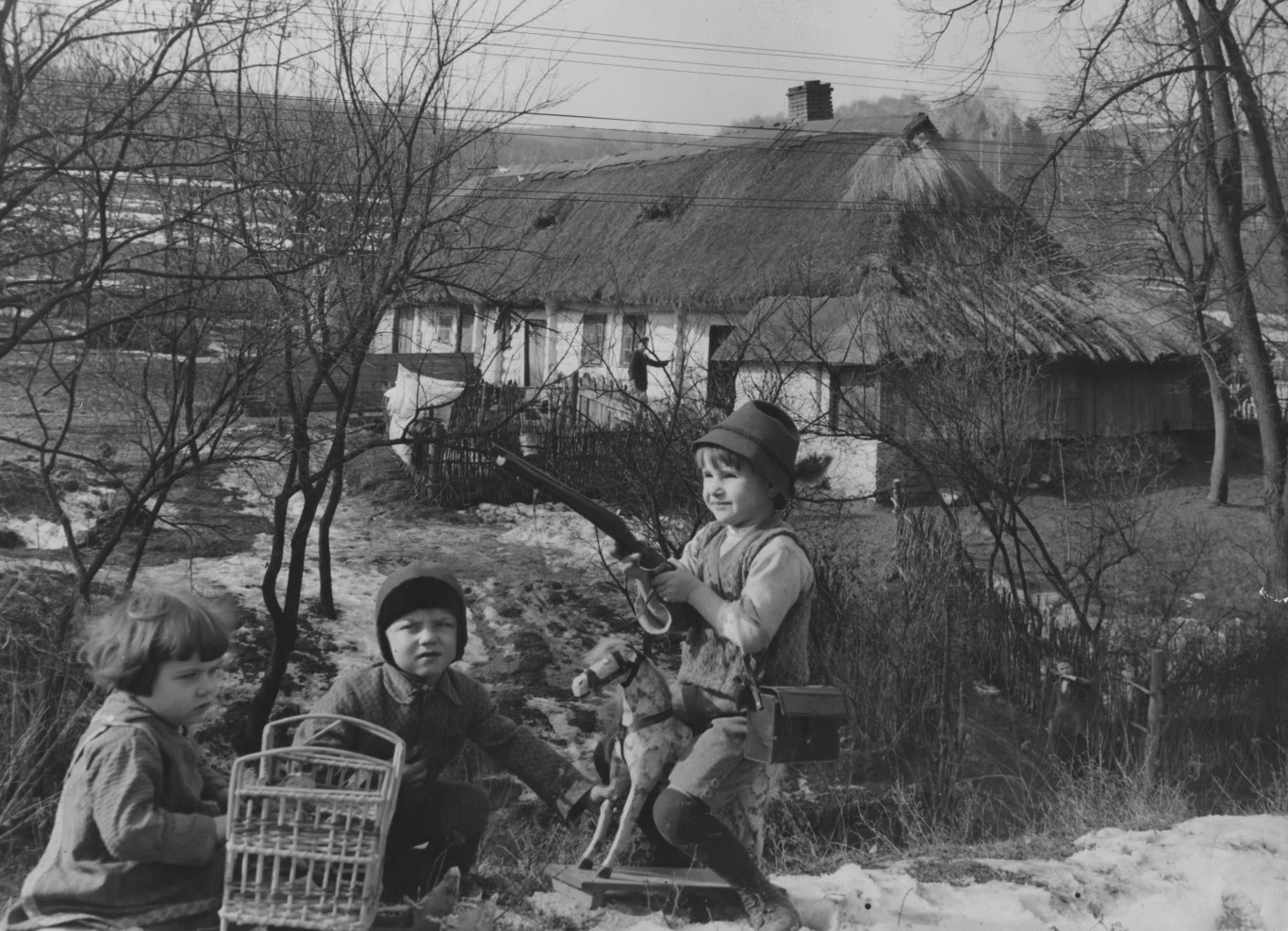 troje dzieci bawi się na tle wiejskiej chaty krytej strzechą; jedno z nich siedzi na drewnianym koniku, trzymając w ręce zabawkową strzelbę, obok stoi koszyk i pluszowy pies; otoczenie tworzą gołe drzewa i resztki śniegu na ziemi