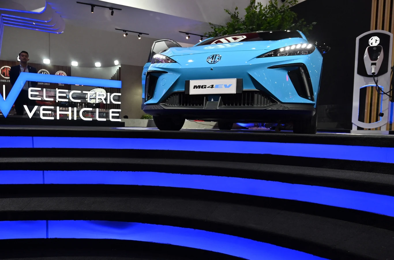 jasnoniebieski samochód elektryczny MG4 EV eksponowany na podwyższeniu obok stacji ładowania, podświetlone napisy 'ELECTRIC VEHICLE' oraz niebieskie światła podkreślające futurystyczny charakter prezentacji