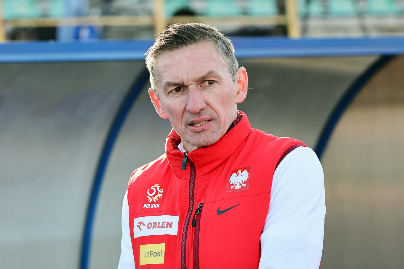 mężczyzna o krótkich, przyciemnionych włosach ubrany w czerwono-białą kamizelkę z logo reprezentacji Polski oraz sponsorów, stojący na tle stadionowych trybun; wyraz twarzy poważny i skupiony
