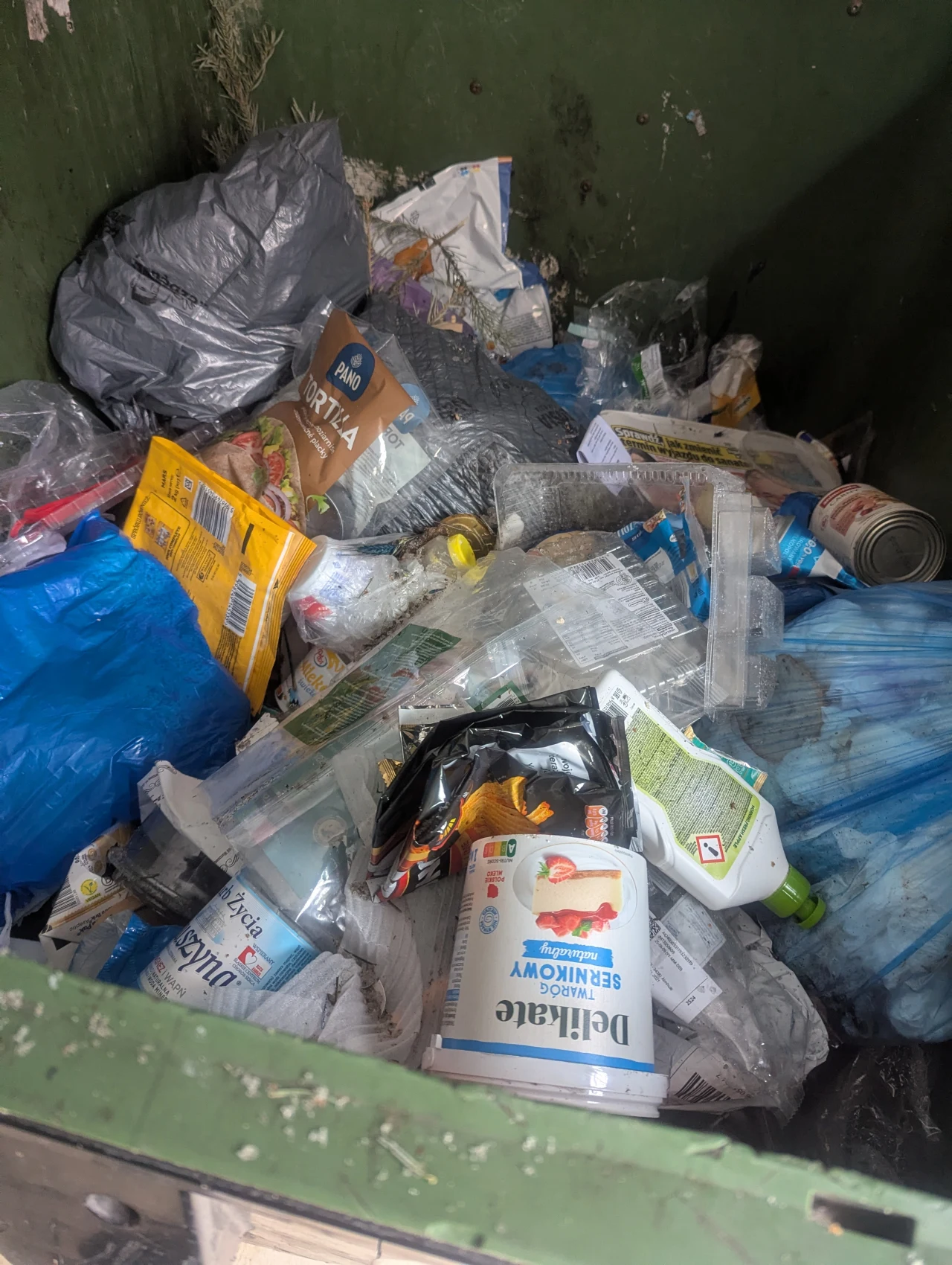 Duża ilość zmieszanych śmieci, głównie plastikowe opakowania po produktach spożywczych, puszki, foliowe torby i zużyte kartony wrzucone do jednego kontenera.
