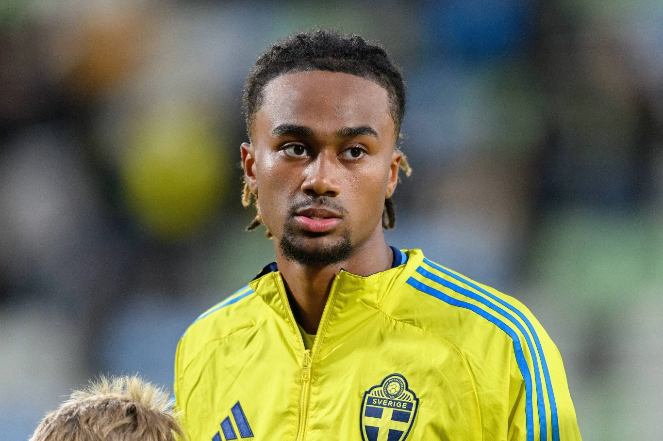 młody mężczyzna w żółtej sportowej kurtce z herbem Szwecji, stojący na tle rozmytego stadionu