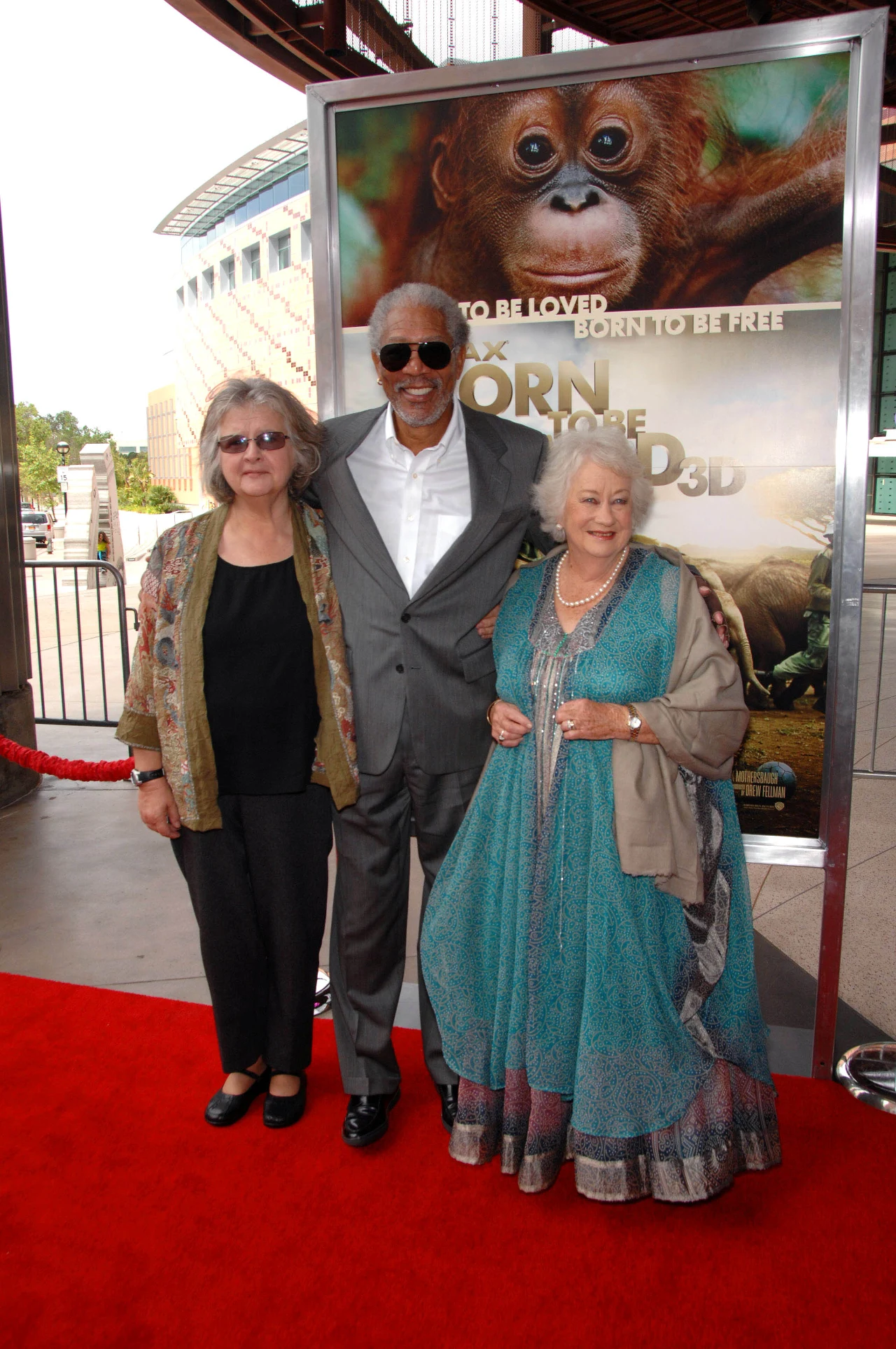 Trzy uśmiechnięte osoby pozują na czerwonym dywanie przy plakacie filmu o orangutanach, w tle widoczna nowoczesna architektura i barierki z czerwonego sznura.