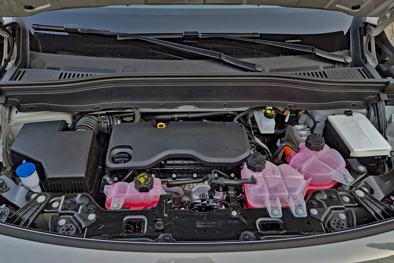 komora silnika samochodu osobowego z widocznym silnikiem benzynowym, zbiornikami płynów eksploatacyjnych oznaczonymi różowym płynem, filtrem powietrza po lewej stronie i akumulatorem po prawej