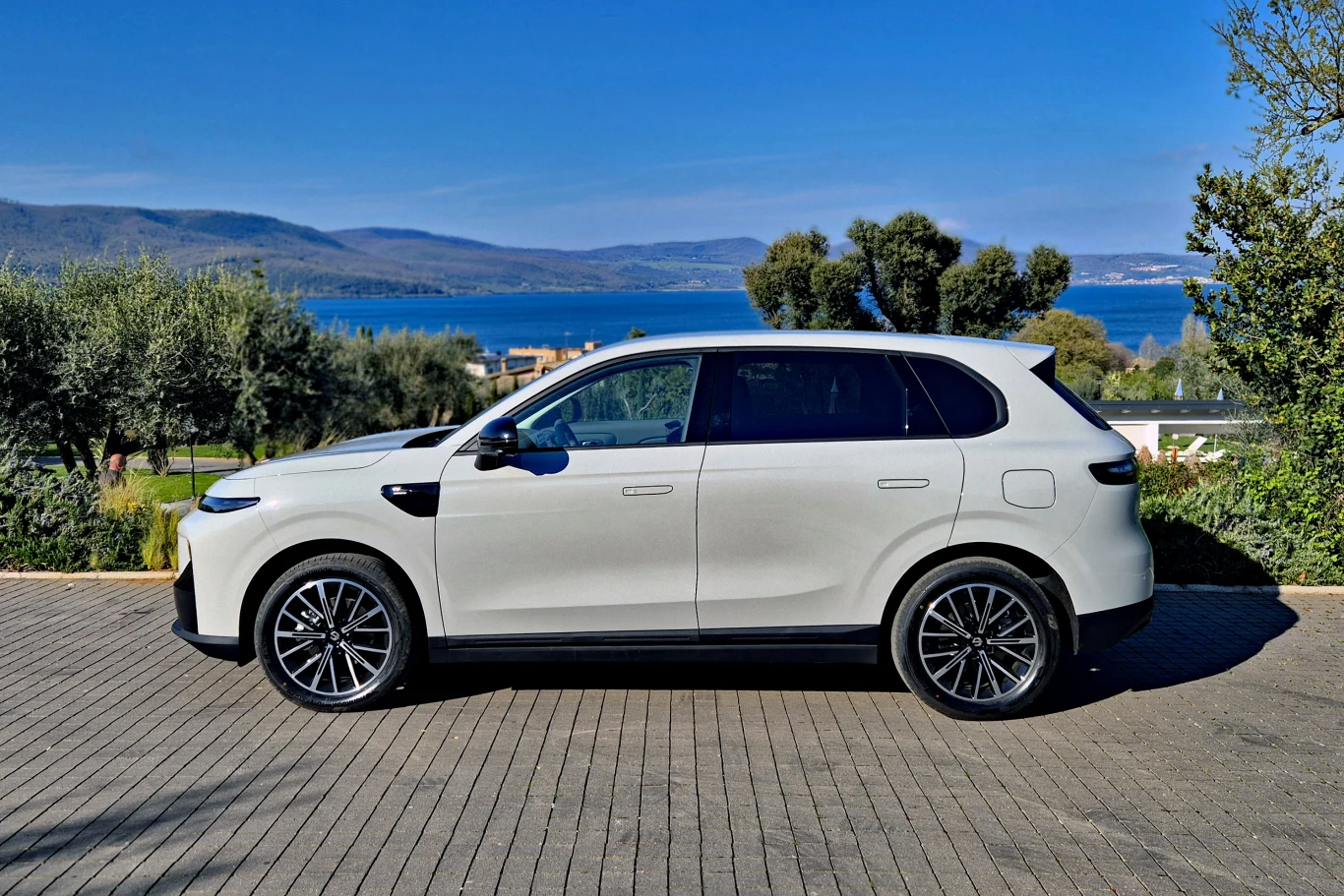 biały, nowoczesny SUV zaparkowany na brukowanym podjeździe, w tle rozpościera się widok na jezioro oraz zielone wzgórza, jasne niebo bez chmur