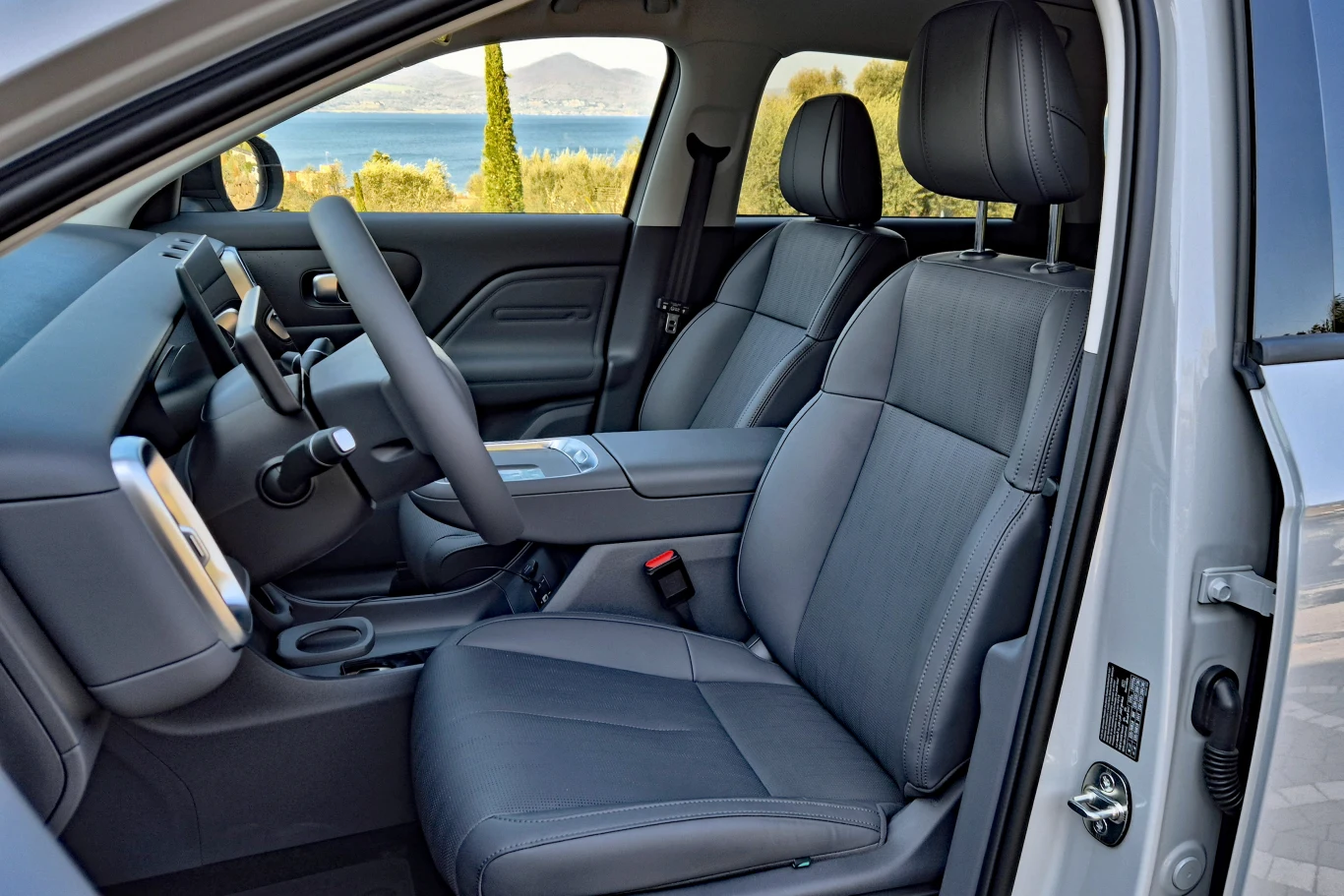 przestronne, nowoczesne wnętrze samochodu z jasnoszarymi, skórzanymi fotelami, minimalistyczna deska rozdzielcza z dużym ekranem, szerokie podłokietniki oraz panoramiczny widok na krajobraz z jeziorem i górami za szybą