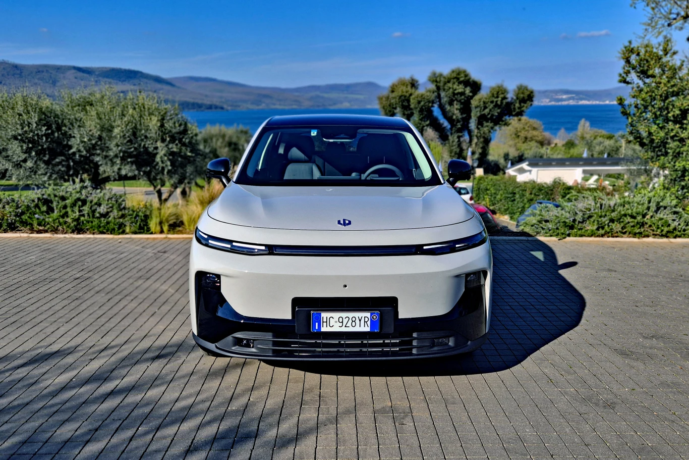 nowoczesny biały samochód elektryczny zaparkowany na brukowanym placu, na tle zielonych drzew i jeziora z górzystym krajobrazem w oddali