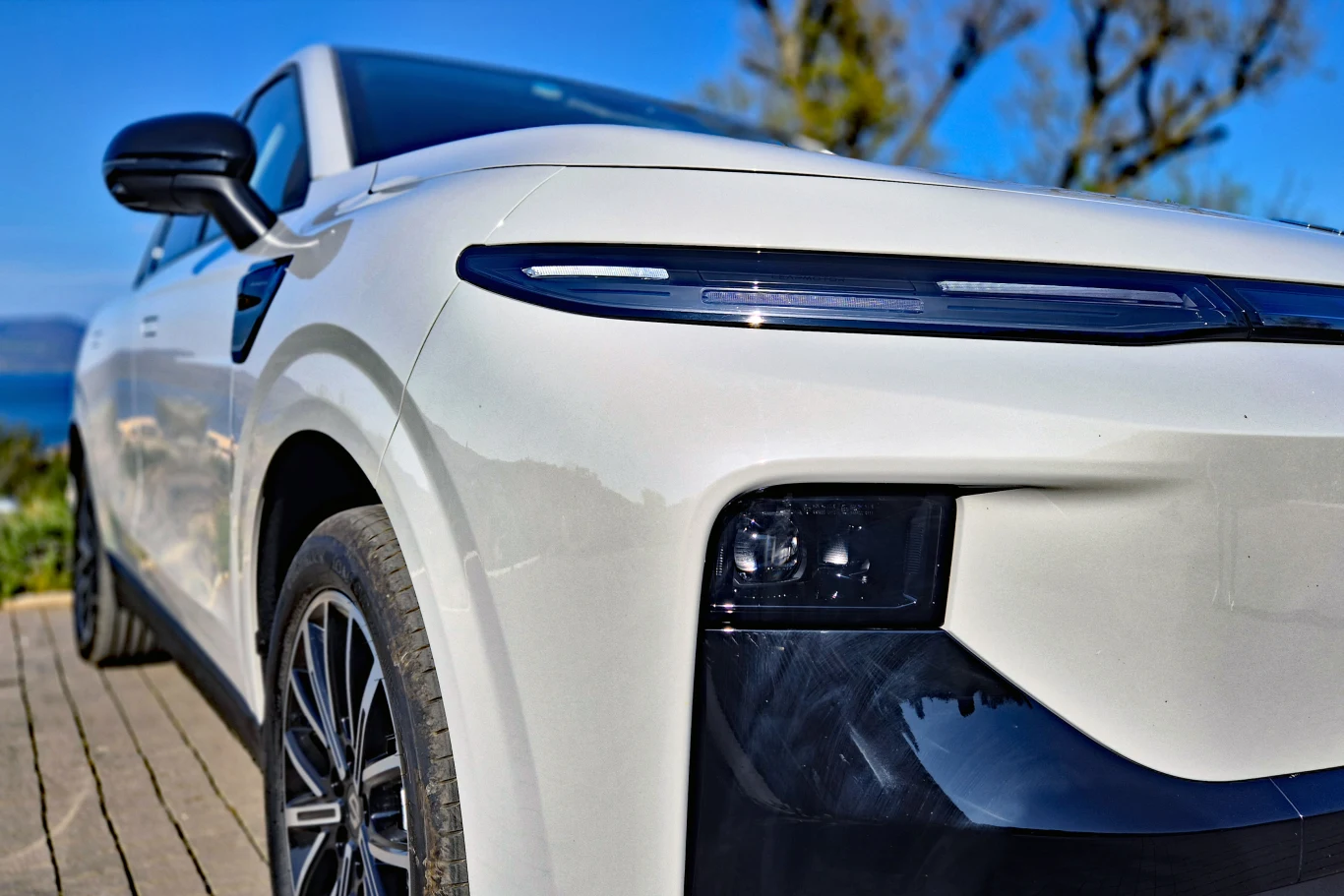 nowoczesny biały samochód typu SUV z wyraźnie zarysowanymi reflektorami LED, masywnym zderzakiem i dużymi kołami, zaparkowany na drewnianym podeście przy jasnym, słonecznym niebie