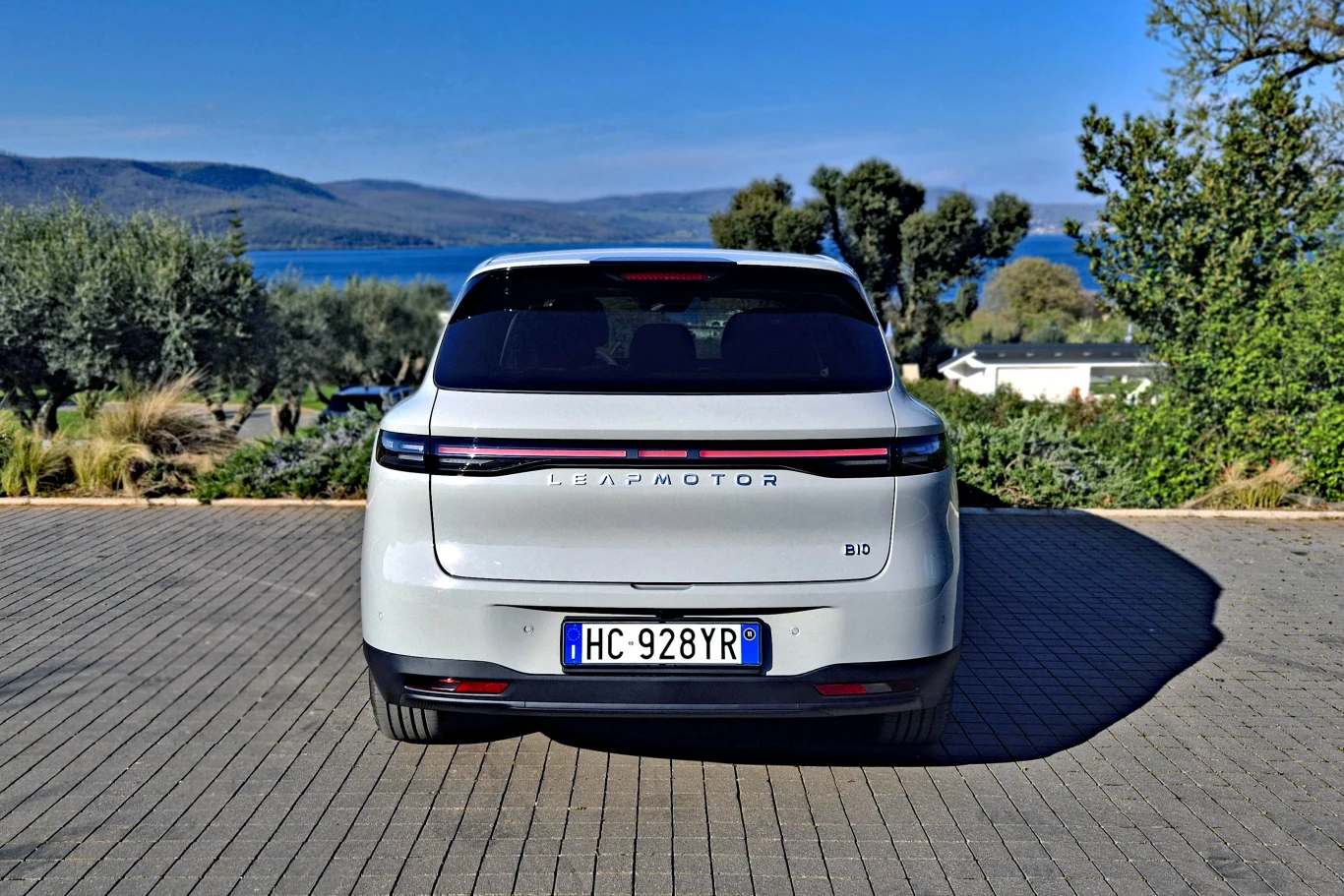 biały samochód marki Leapmotor B10 widoczny od tyłu, zaparkowany na brukowanym podjeździe, w tle jezioro oraz drzewa i wzniesienia