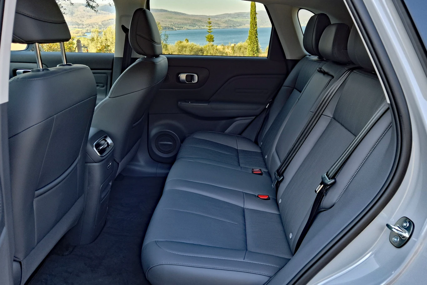 tylna kanapa samochodu w ciemnej tapicerce, z trzema zagłówkami, pasami bezpieczeństwa i przestronną przestrzenią na nogi, przy bocznym oknie widoczny krajobraz z jeziorem i wzgórzami