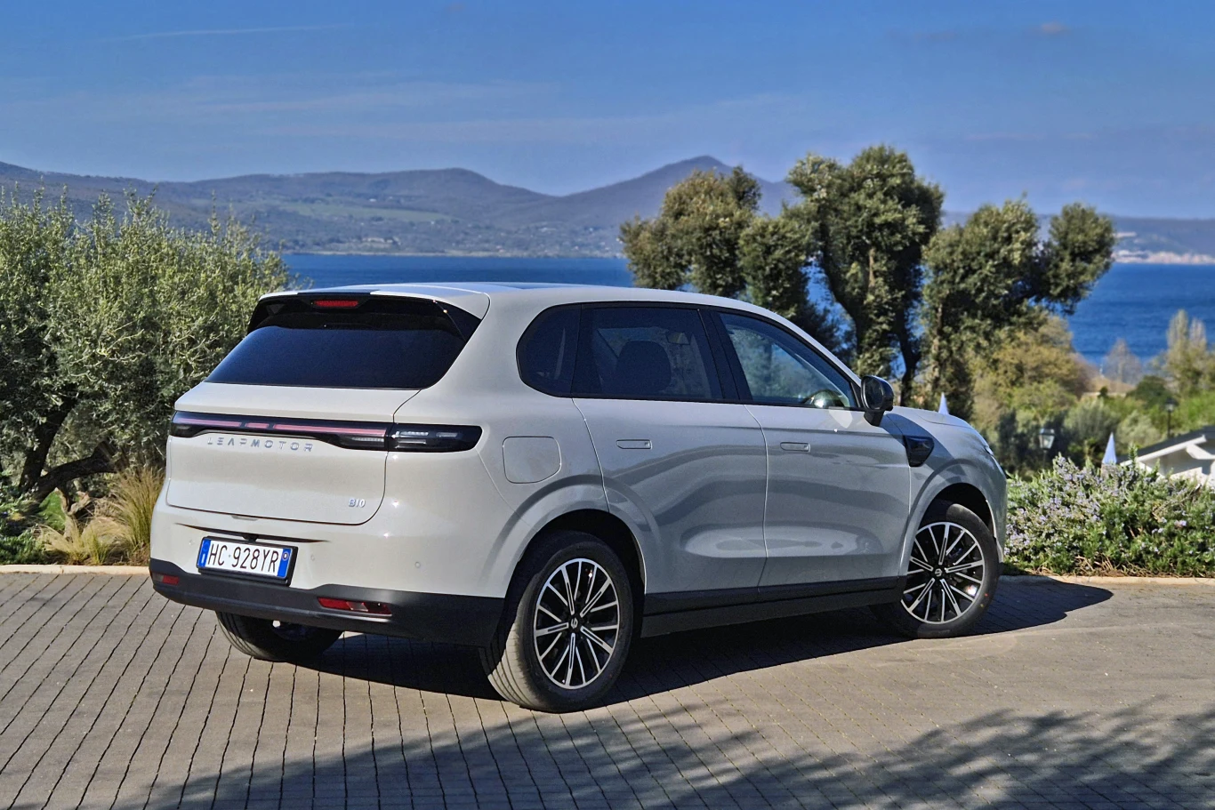 jasny SUV marki Seres 5 zaparkowany na brukowanym podjeździe, otoczony zielenią i drzewami, w tle widoczny krajobraz górski oraz fragment niebieskiego jeziora lub morza