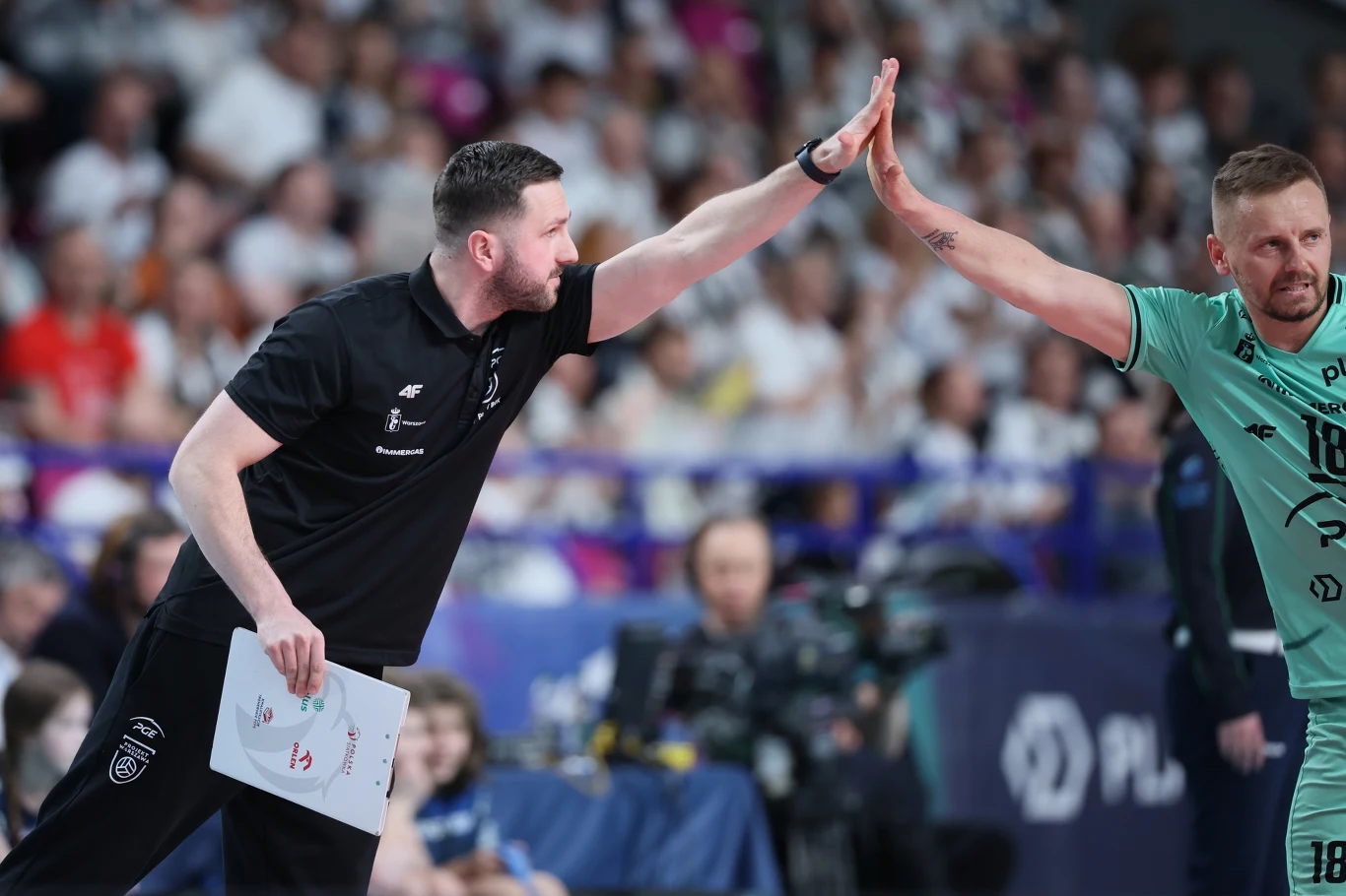 trener drużyny siatkarskiej w czarnym stroju przybija piątkę zawodnikowi ubranym w zielony strój sportowy, tłum kibiców w tle