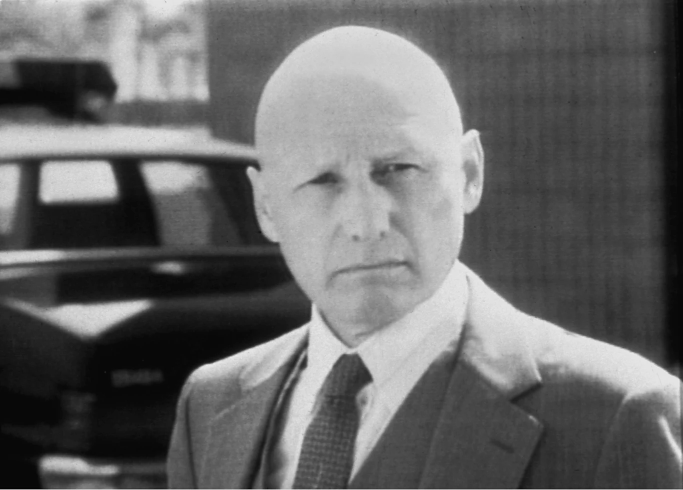 Łysy mężczyzna w garniturze i krawacie patrzy poważnie w stronę obiektywu; w tle rozmyty czarny samochód.