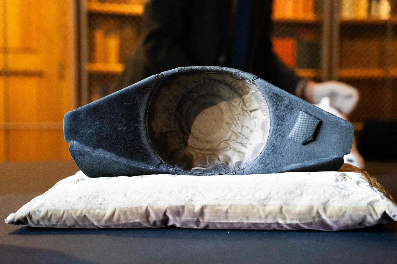 Starożytna korona z ciemnego materiału z wyraźnie widocznym, wytartym wnętrzem, umieszczona na haftowanej poduszce; w tle półki z książkami oraz zamazana postać w rękawiczkach.