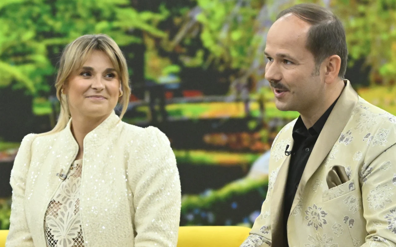 Sławomir i Kajra Dwoje elegancko ubranych ludzi siedzi obok siebie na żółtej sofie; kobieta uśmiecha się łagodnie, a mężczyzna mówi, gestykulując dłonią, w tle rozmyty motyw roślinno-parkowy.