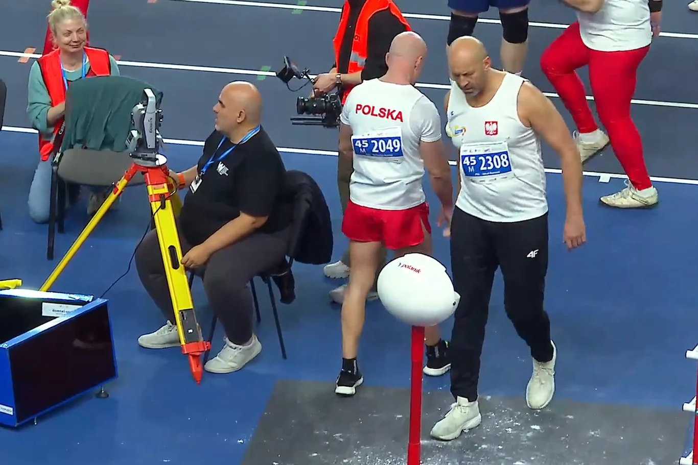 dwóch lekkoatletów w strojach reprezentacji Polski przygotowuje się do udziału w zawodach sportowych na hali, jeden z zawodników stoi przy stojaku z białą kulą do pchnięcia kulą, w tle sędziowie i sprzęt pomiarowy