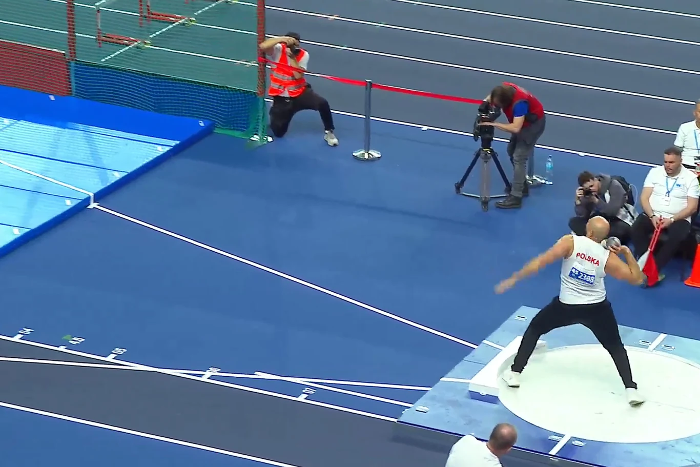 zawodnik w białej koszulce z napisem 'POLAND' przygotowuje się do rzutu kulą na arenie lekkoatletycznej, podczas gdy kilku fotografów i sędziowie obserwują jego występ