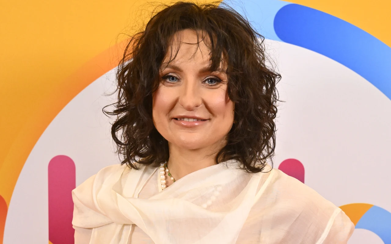 kobieta z ciemnymi, kręconymi włosami i delikatnym makijażem, ubrana w jasną bluzkę z perłowym naszyjnikiem, stoi na tle kolorowej, graficznej ściany z widocznymi okręgami w odcieniach niebieskiego, różowego i pomarańczowego