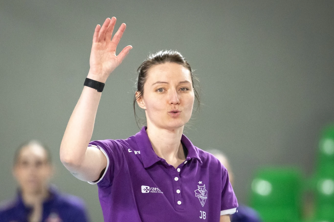 Kobieta w fioletowej koszulce sportowej z uniesioną ręką, stojąca na hali sportowej z zielonymi krzesłami w tle.