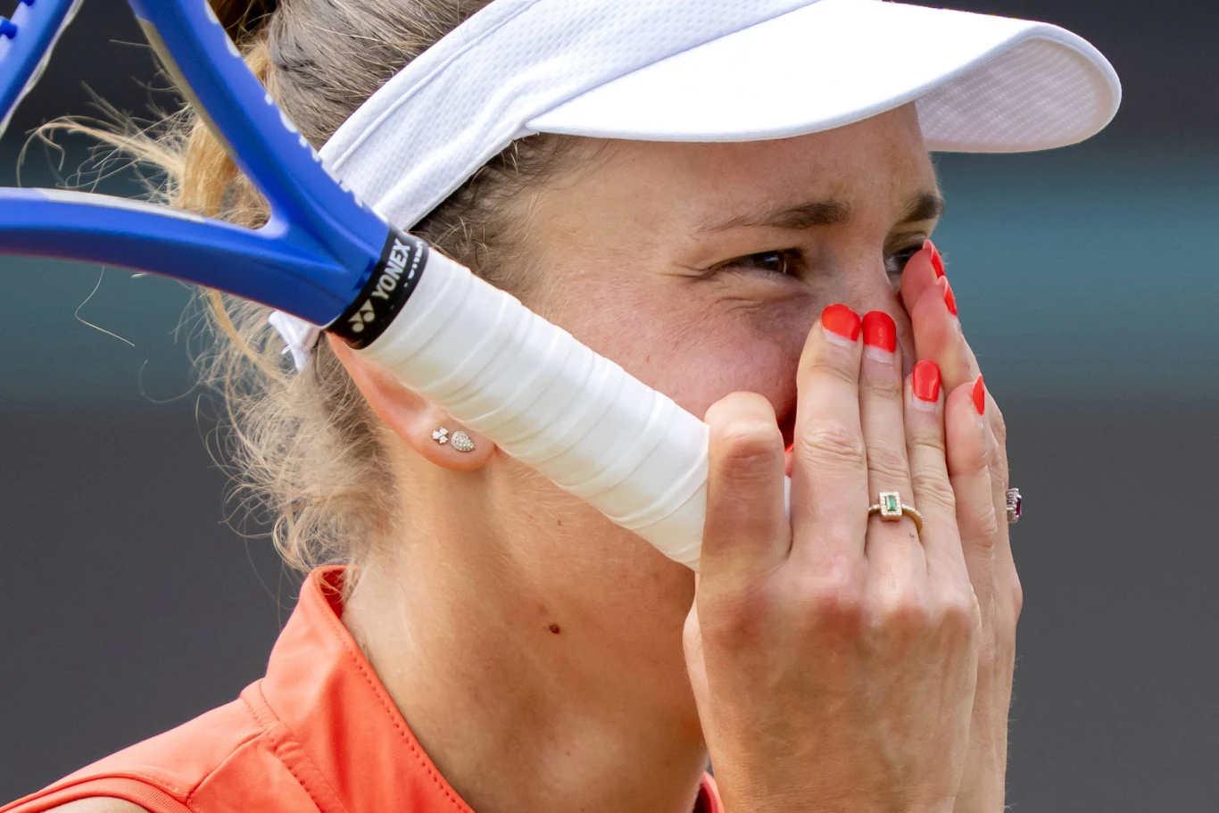 Zbliżenie na tenisistkę w sportowej czapeczce i pomarańczowej koszulce, trzymającą rakietę tenisową oraz zakrywającą usta dłońmi z czerwonym lakierem do paznokci; na twarzy widoczny wyraz silnych emocji.
