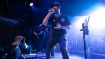 wokalista w sportowej koszulce energetycznie śpiewa do mikrofonu na jasno oświetlonej scenie, w tle widoczni gitarzyści oraz fotograf po lewej stronie rejestrujący występ