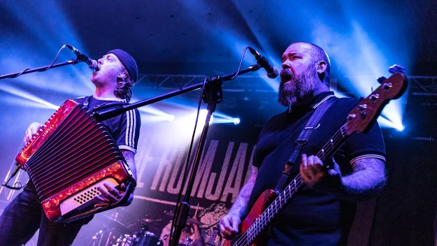 Dwóch muzyków wykonuje utwór na scenie oświetlonej niebieskim światłem; jeden gra na akordeonie i śpiewa do mikrofonu, drugi gra na gitarze basowej i również śpiewa; w tle widoczne są elementy perkusji i nazwa zespołu na banerze.