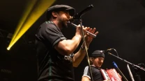 Dwóch muzyków występuje na scenie; jeden gra na flecie poprzecznym i śpiewa do mikrofonu, drugi gra na czerwonym akordeonie, obaj są skupieni na grze, oświetleni światłem scenicznym.