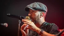 mężczyzna w czapce z daszkiem i sportowej koszulce śpiewa do mikrofonu na scenie, z intensywnym światłem skierowanym na niego
