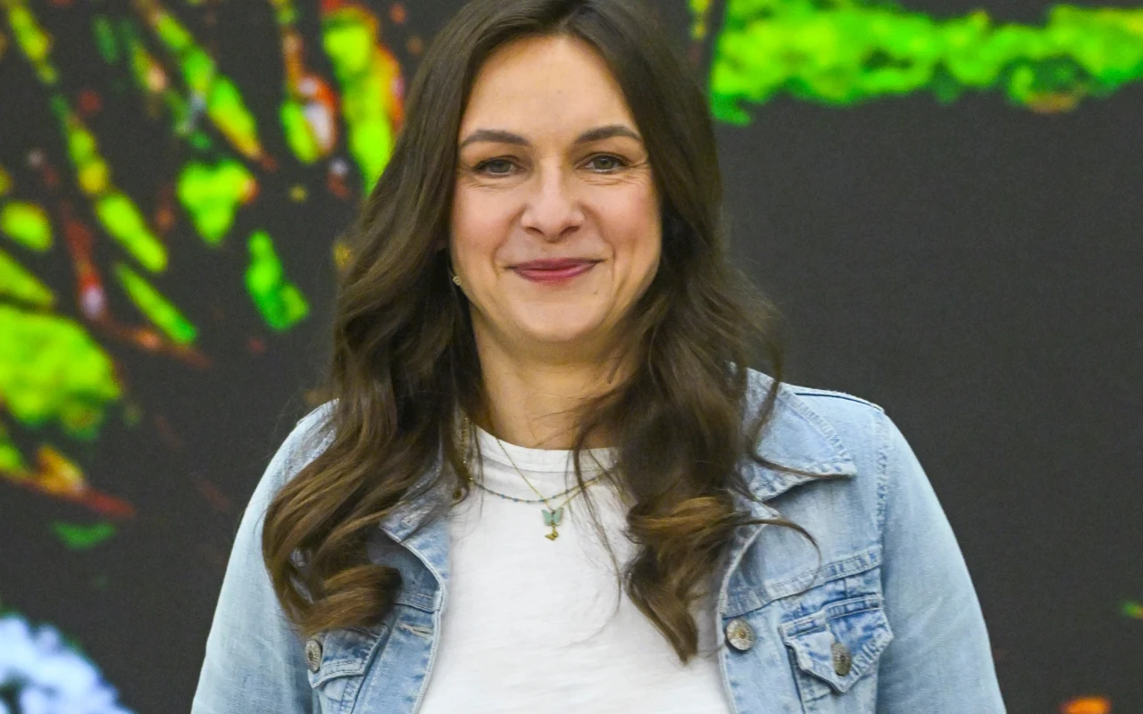 Joanna Drożyńska Joanna Drożyńska uśmiecha się w "halo tu polsat", obrana w jeansową kurtkę i białą koszulkę, na tle zielonego, rozmytego tła.