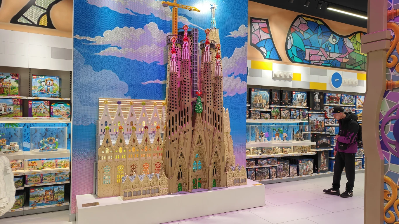 duża makieta Sagrada Familia zbudowana z klocków Lego, ustawiona na tle kolorowej mozaiki ściennej z motywem nieba i chmur, w otoczeniu licznych półek z różnorodnymi zestawami Lego, obok której stoi osoba oglądająca ekspozycję