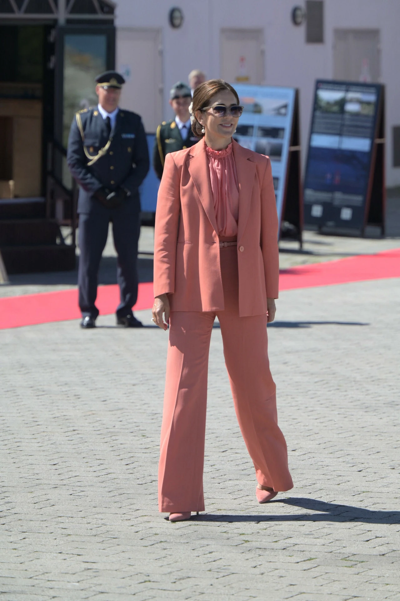 kobieta w eleganckim, łososiowym garniturze stoi na wybrukowanym placu przy czerwonym dywanie; w tle dwóch mężczyzn w mundurach oraz tablice z prezentacjami