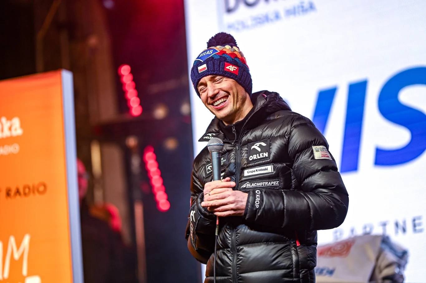 Uśmiechnięty mężczyzna w czarnej, zimowej kurtce i czapce z widocznymi logotypami sponsorów stoi na scenie, trzymając w rękach mikrofon, w tle oświetlona scena oraz ekrany z reklamami i napisami.