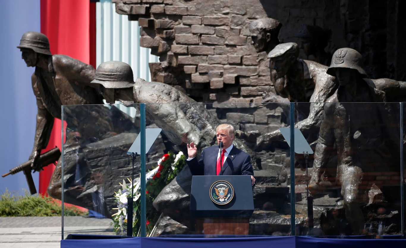 Donald Trump stoi za mównicą z herbem Stanów Zjednoczonych, gestykulując podczas przemówienia przed monumentalnym pomnikiem przedstawiającym żołnierzy w dynamicznych pozach, z widocznymi barierami ochronnymi ze szkła wokół mównicy.